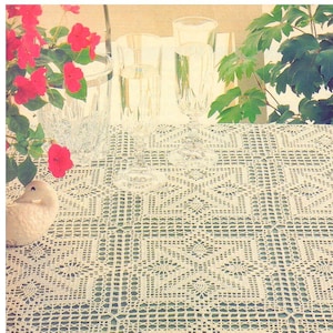 May include: A white crocheted tablecloth with a detailed pattern, set with champagne flutes filled with a bubbly beverage. Red flowers in a clear vase and a decorative duck figurine add to the scene. Green foliage frames the background.