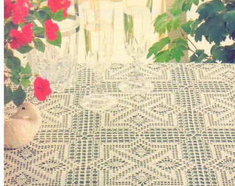 May include: A cream-colored crocheted tablecloth featuring a repeating square geometric pattern with intricate floral-like centers. The textile is displayed with two clear glass flutes, a crystal vase, a small ceramic duck, and green foliage.