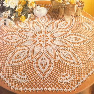 Puede incluir: Un tapete de ganchillo redondo y blanco que presenta un intrincado patrón floral radiante con motivos en forma de hoja y un delicado borde de malla calada. El diseño se centra en un pequeño núcleo en forma de estrella que se expande en segmentos.