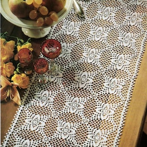 Puede incluir: Un camino de mesa de crochet blanco con un delicado patrón floral. El camino de mesa está colocado sobre una mesa de madera con un tazón de fruta y vasos.