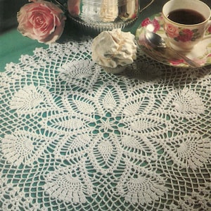 May include: White crocheted doily with a delicate floral pattern, perfect for a table setting. The doily is laid on a green surface with a silver sugar bowl, a cup of coffee, and a pink rose.