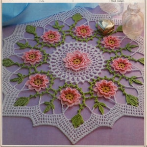 Peut inclure: Une dentelle crochetée blanche avec un motif floral délicat. La dentelle présente des fleurs roses avec des centres jaunes et des feuilles vertes. La dentelle mesure environ 30 cm de diamètre.