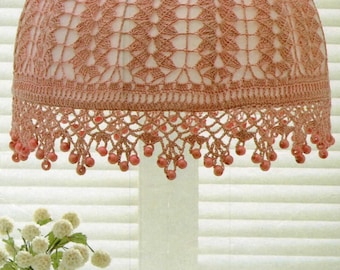 May include: A pink crocheted lampshade with a beaded trim. The lampshade is hanging from a ceiling fixture. The lampshade is in front of a white window blind.