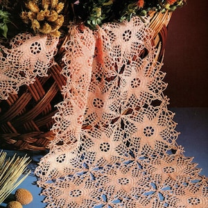 May include: A peach-coloured, open-weave crochet table runner with a floral pattern. The runner is draped over a blue surface, with a wicker basket and dried flowers in the background. The runner's dimensions are approximately 152 cm long.