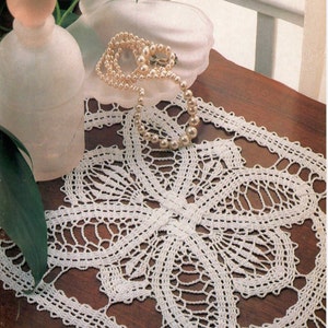 May include: A white, crocheted doily with a floral design is displayed on a wooden surface. A pearl necklace and a frosted glass bottle with a round stopper are also present. The doily's intricate openwork pattern is visible.