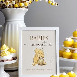 Pu&ograve; includere: Un cartello incorniciato di bianco con il testo "Babies are sweet" e un cartone animato di Winnie the Pooh che tiene un barattolo di miele. Il cartello &egrave; su un tavolo con cupcake gialli.