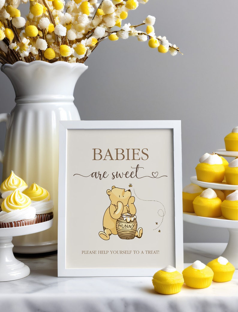 Pu&ograve; includere: Un cartello incorniciato di bianco con il testo "Babies are sweet" e un cartone animato di Winnie the Pooh che tiene un barattolo di miele. Il cartello &egrave; su un tavolo con cupcake gialli.