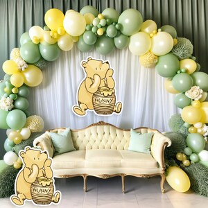 May include: A Winnie the Pooh themed party decoration featuring a green and yellow balloon arch, a white backdrop, and a cream-coloured sofa with green cushions. Two Winnie the Pooh cutouts are positioned on either side of the sofa, each holding a honey pot labelled "Hunny".