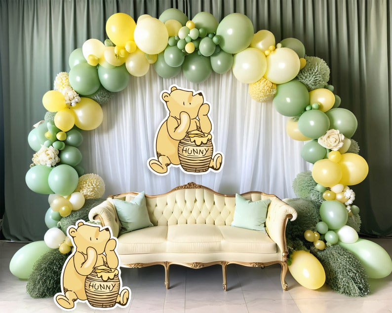 May include: A Winnie the Pooh themed party decoration featuring a green and yellow balloon arch, a white backdrop, and a cream-coloured sofa with green cushions. Two Winnie the Pooh cutouts are positioned on either side of the sofa, each holding a honey pot labelled "Hunny".