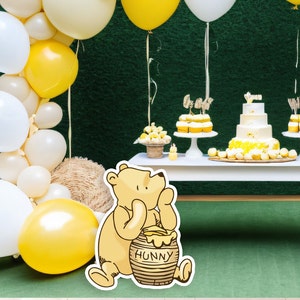 May include: A Winnie the Pooh themed baby shower with a white table decorated with yellow and white balloons, cupcakes, and a cake. A Winnie the Pooh character holding a honey pot with the word "Hunny" on it is in the foreground.