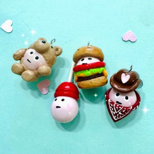 May include: A collection of four handmade clay charms. The charms include a brown bear, a burger, a snowman with a red hat, and a cowboy with a brown hat and red bandana. Each charm has a small metal loop for hanging.