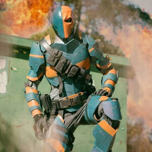 May include: A blue and orange superhero costume with a helmet and a detailed design. The costume is worn by a person standing in front of a green skip with smoke and fire in the background.