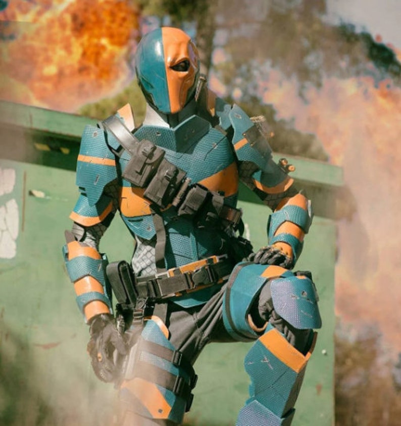 May include: A blue and orange superhero costume with a helmet and a detailed design. The costume is worn by a person standing in front of a green skip with smoke and fire in the background.