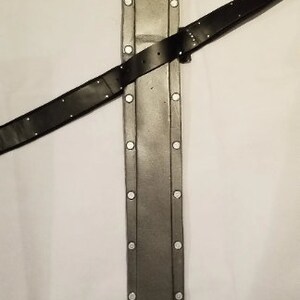 May include: A grey leather scabbard with a black leather strap. The scabbard has a pointed tip and a rounded top. The strap is attached to the scabbard with metal rivets.