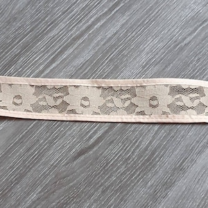 Könnte beinhalten: Ein beigefarbener Spitzenbesatz mit einem Blumenmuster. Der Besatz hat einen gewellten Rand und ist mit einem hellbeigen Faden vernäht.