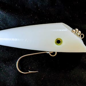 May include: A white fishing lure with a streamlined design. It features a silver hook and a small, green eye. The lure is designed to attract fish and is likely used for recreational fishing.