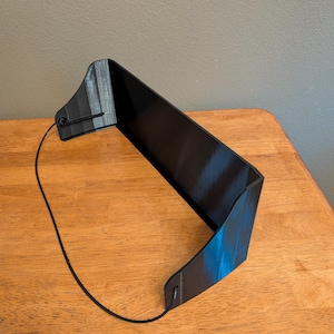 May include: A black, U-shaped plastic visor or shield with a smooth, reflective surface. The object features two side panels connected by a central bridge, with a thin, black elastic cord attached to the side holes for securing the item in place.