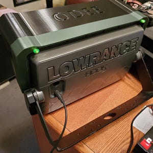 Może przedstawiać: Szary echosonda Lowrance HOOK z czarnym wspornikiem, zamontowany na drewnianej powierzchni. Urządzenie ma widoczne słowo "LOWRANCE". Na g&oacute;rze urządzenia wytłoczono słowo "ODEE". Widoczne są zielone światła.