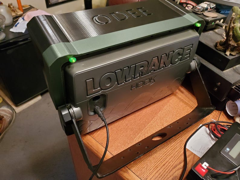 Może przedstawiać: Szary echosonda Lowrance HOOK z czarnym wspornikiem, zamontowany na drewnianej powierzchni. Urządzenie ma widoczne słowo "LOWRANCE". Na g&oacute;rze urządzenia wytłoczono słowo "ODEE". Widoczne są zielone światła.
