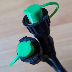 May include: Close-up of two black plastic connectors with green caps. The caps are attached to the connectors with a green flexible tether. The connectors have a red seal and are on a light brown wooden surface.