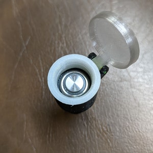 May include: A white plastic battery holder with a black base and a clear plastic lid. The lid is open and a silver battery is visible inside.