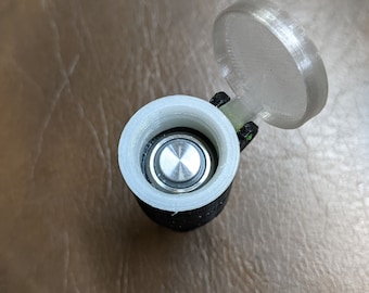 May include: A white plastic battery holder with a black base and a clear plastic lid. The lid is open and a silver battery is visible inside.