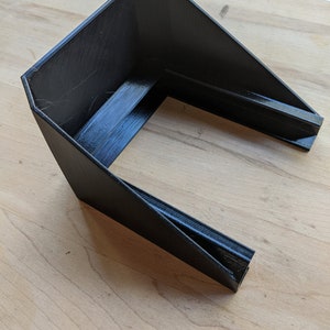 May include: A black plastic 3D printed object with a rectangular opening. The object is designed to be used as a phone stand.