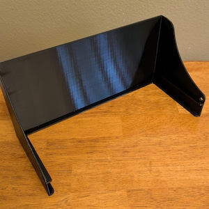 May include: A black, 3D-printed object with a rectangular top and angled sides. The object is sitting on a wooden surface. The design features a curved front edge and two small holes on the side.