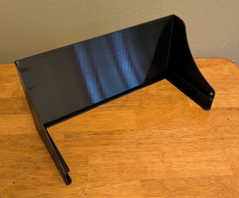 May include: A black, 3D-printed object with a rectangular top and angled sides. The object is sitting on a wooden surface. The design features a curved front edge and two small holes on the side.