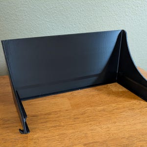 Op de afbeelding: Een zwarte, schuine displaystandaard met een rechthoekige achterkant en gebogen zijkanten. De standaard heeft een kleine haak aan de onderkant en is gemaakt van een mat materiaal. Het is ontworpen om items rechtop te houden.