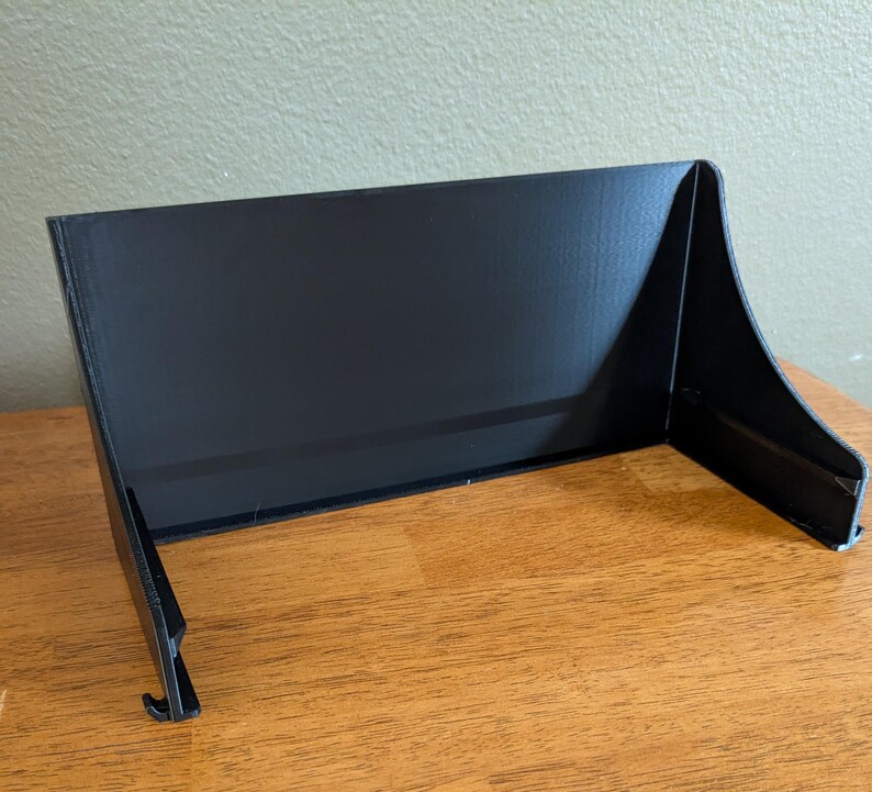 Op de afbeelding: Een zwarte, schuine displaystandaard met een rechthoekige achterkant en gebogen zijkanten. De standaard heeft een kleine haak aan de onderkant en is gemaakt van een mat materiaal. Het is ontworpen om items rechtop te houden.