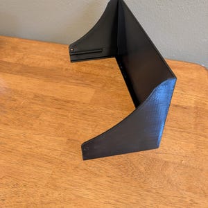 May include: A black, angular object with a unique shape, possibly a shelf or display stand. The object is made of a solid material and has a textured surface. It is positioned on a wooden surface, with a neutral-colored wall in the background.