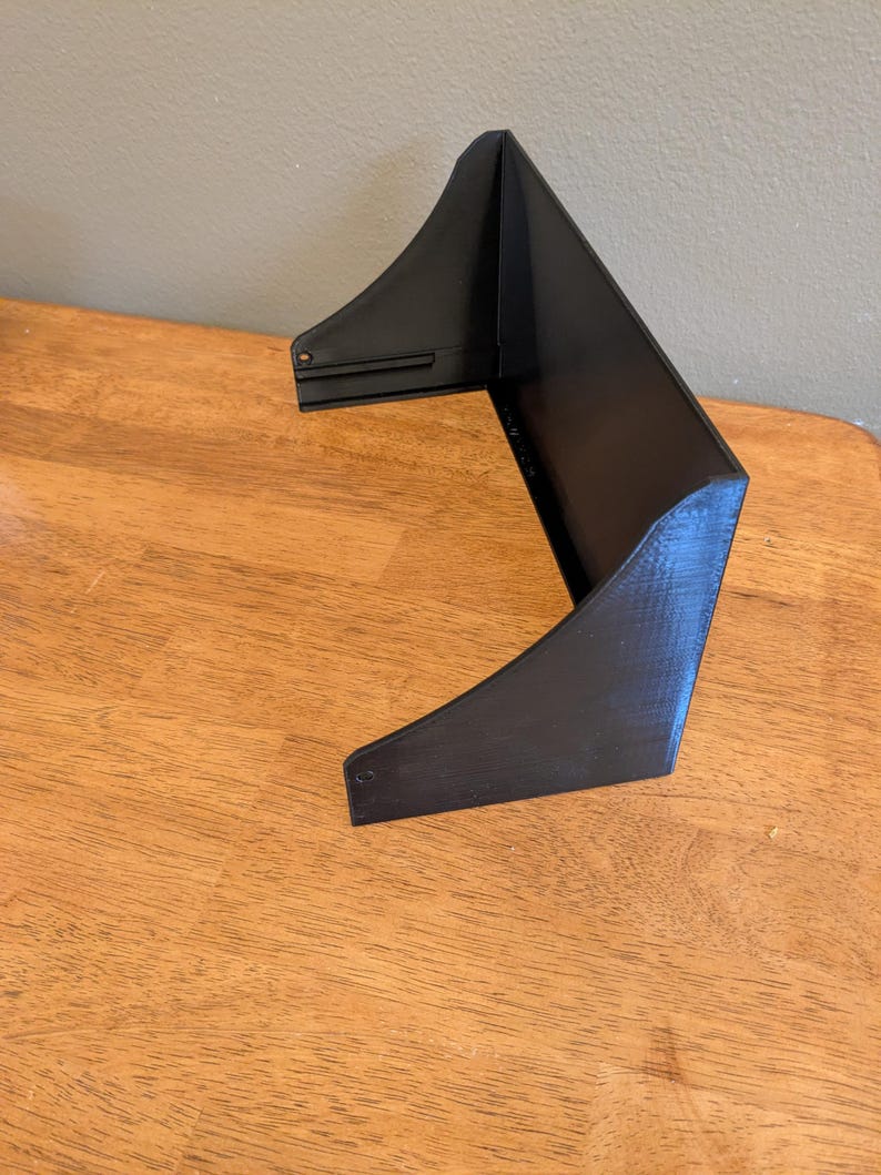 May include: A black, angular object with a unique shape, possibly a shelf or display stand. The object is made of a solid material and has a textured surface. It is positioned on a wooden surface, with a neutral-colored wall in the background.