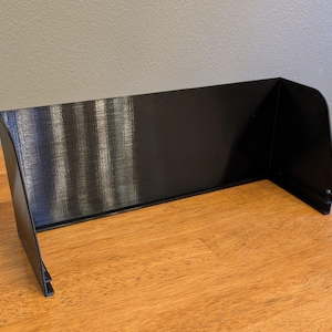 May include: A black, rectangular visor with a curved top and side panels. The visor is made of a solid, opaque material and is designed to block sunlight. It is placed on a wooden surface.