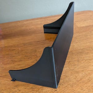 Op de afbeelding: Een zwarte, schuine displaystandaard met een gebogen ontwerp. De standaard is gemaakt van een solide materiaal en heeft een platte basis. De standaard is ontworpen om een item rechtop te houden.