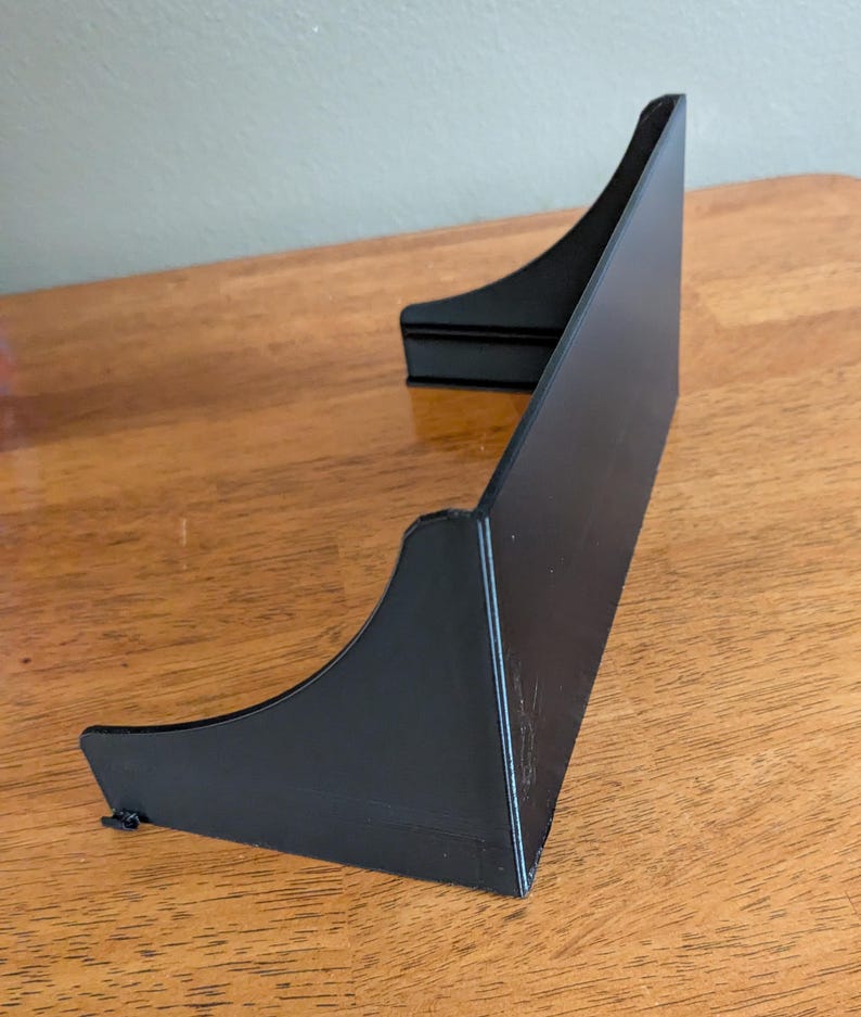 Op de afbeelding: Een zwarte, schuine displaystandaard met een gebogen ontwerp. De standaard is gemaakt van een solide materiaal en heeft een platte basis. De standaard is ontworpen om een item rechtop te houden.