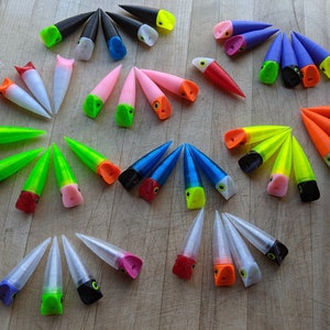 May include: A collection of colorful, cone-shaped fishing lures in various colors, including black, white, pink, green, blue, orange, and yellow. Each lure has a contrasting tip and a small eye detail, arranged on a wooden surface.