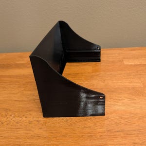May include: A black, 3D-printed shelf with a curved design. The shelf has a rectangular base and two side supports that curve upwards. The shelf is made of a solid black material and sits on a wooden surface.