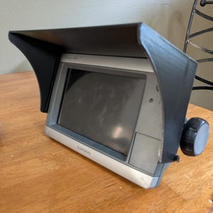 Op de afbeelding: Een Garmin GPS-apparaat met een zwarte zonneklep. Het apparaat heeft een zilveren rand en een scherm. Aan de rechterkant zit een zwarte knop. Het apparaat staat op een houten oppervlak.