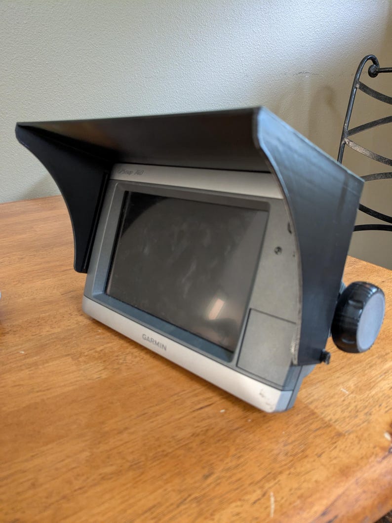 Op de afbeelding: Een Garmin GPS-apparaat met een zwarte zonneklep. Het apparaat heeft een zilveren rand en een scherm. Aan de rechterkant zit een zwarte knop. Het apparaat staat op een houten oppervlak.