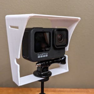 May include: A white 3D printed GoPro camera mount with a sunshade. The GoPro camera is black and has the GoPro logo on the front.
