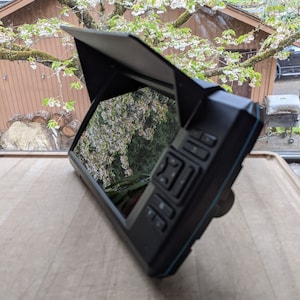 May include: A black and blue portable fish finder with a screen that is displaying a view of a tree with white flowers. The fish finder has a sunshade and multiple buttons.