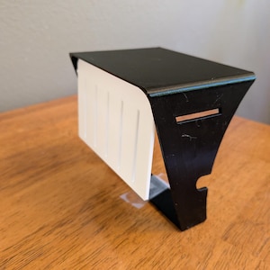 May include: A black metal shelf bracket with a flat top surface and a vertical side panel featuring a white rectangular insert with four vertical grooves. The bracket has a cutout on the side and a horizontal slot near the top edge.