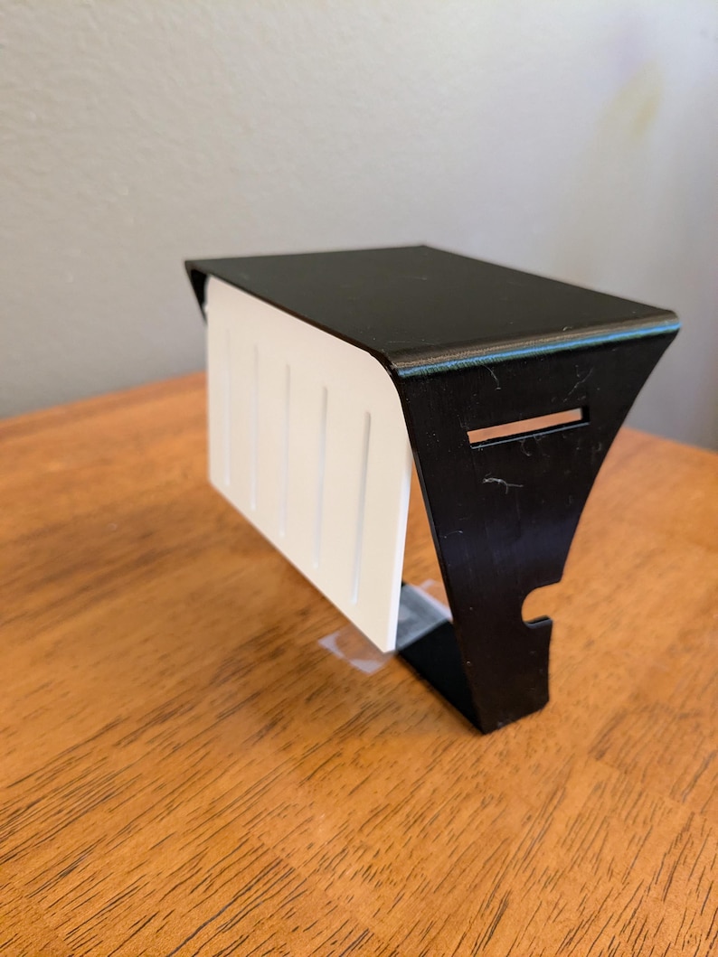 May include: A black metal shelf bracket with a flat top surface and a vertical side panel featuring a white rectangular insert with four vertical grooves. The bracket has a cutout on the side and a horizontal slot near the top edge.