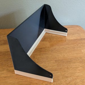 May include: A black and white angled shelf. The shelf has a black, angled top and sides with a white base. The shelf is made of a solid material and has a smooth finish. The shelf is designed to be mounted on a wall.