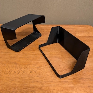 May include: Two black plastic monitor hoods, designed to block glare from overhead lights. The hoods are angled to fit over a computer monitor.