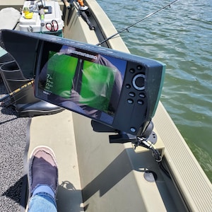 May include: A Humminbird fish finder mounted on a boat. The screen displays green and white sonar data. The boat is olive green, and a fishing rod is visible. A person's foot in blue jeans and a shoe is in the foreground.