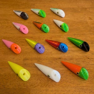 May include: A collection of colorful fishing lures in various designs. The lures feature a pointed shape with a hole at the base and are painted in shades of pink, orange, green, yellow, white, and black. Some have stripes or contrasting color sections.