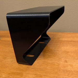 May include: A black, angled shelf with a curved design. The shelf has a flat top and a series of holes along the bottom edge. The shelf is made of a solid material and is designed to be mounted.