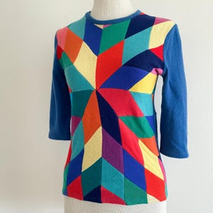 Op de afbeelding: Een blauwe trui met een geometrisch patroon in verschillende kleuren en driekwart mouwen.